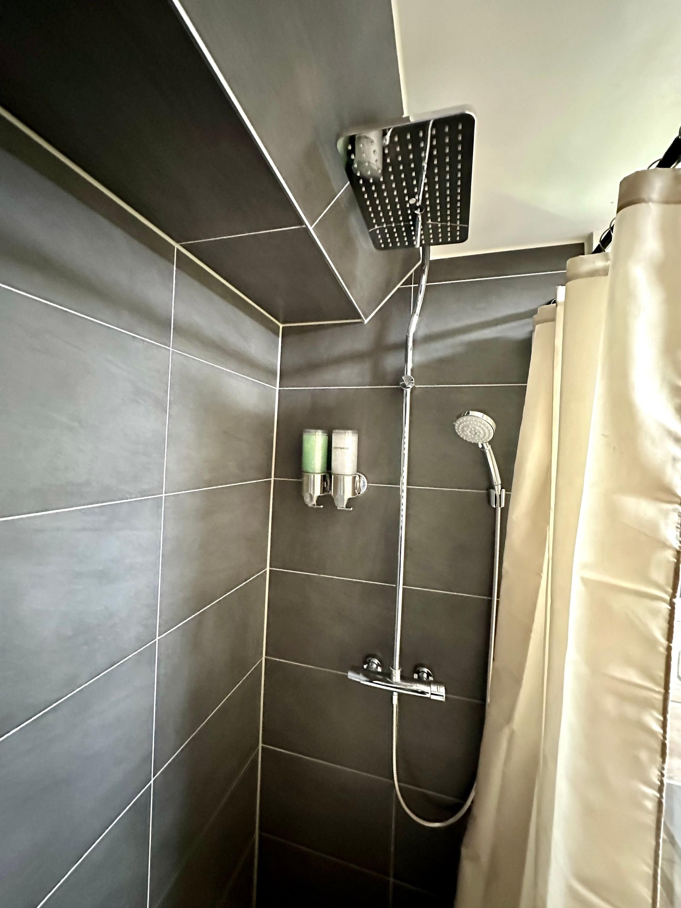 A shower area with dark tiles, a handheld showerhead, a wall-mounted rain showerhead, and a beige shower curtain.