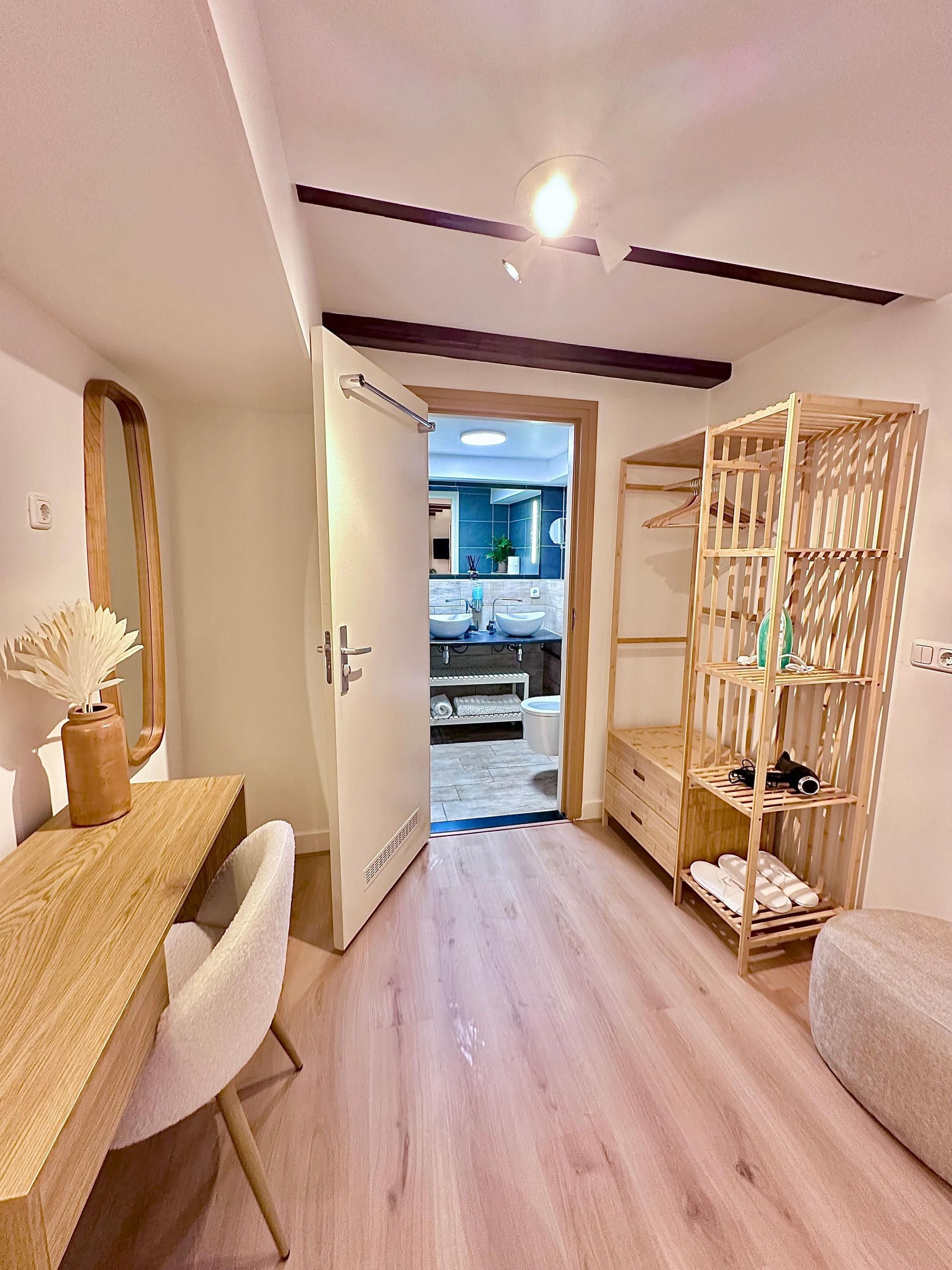 A modern bedroom with a wooden desk, a mirror, a wardrobe, and a view into the bathroom.