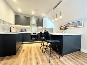 A modern kitchen with dark cabinetry, a wooden floor, and a bar area with two stools. - Windermere Apartments - Long Term Rental Available (Windermere)