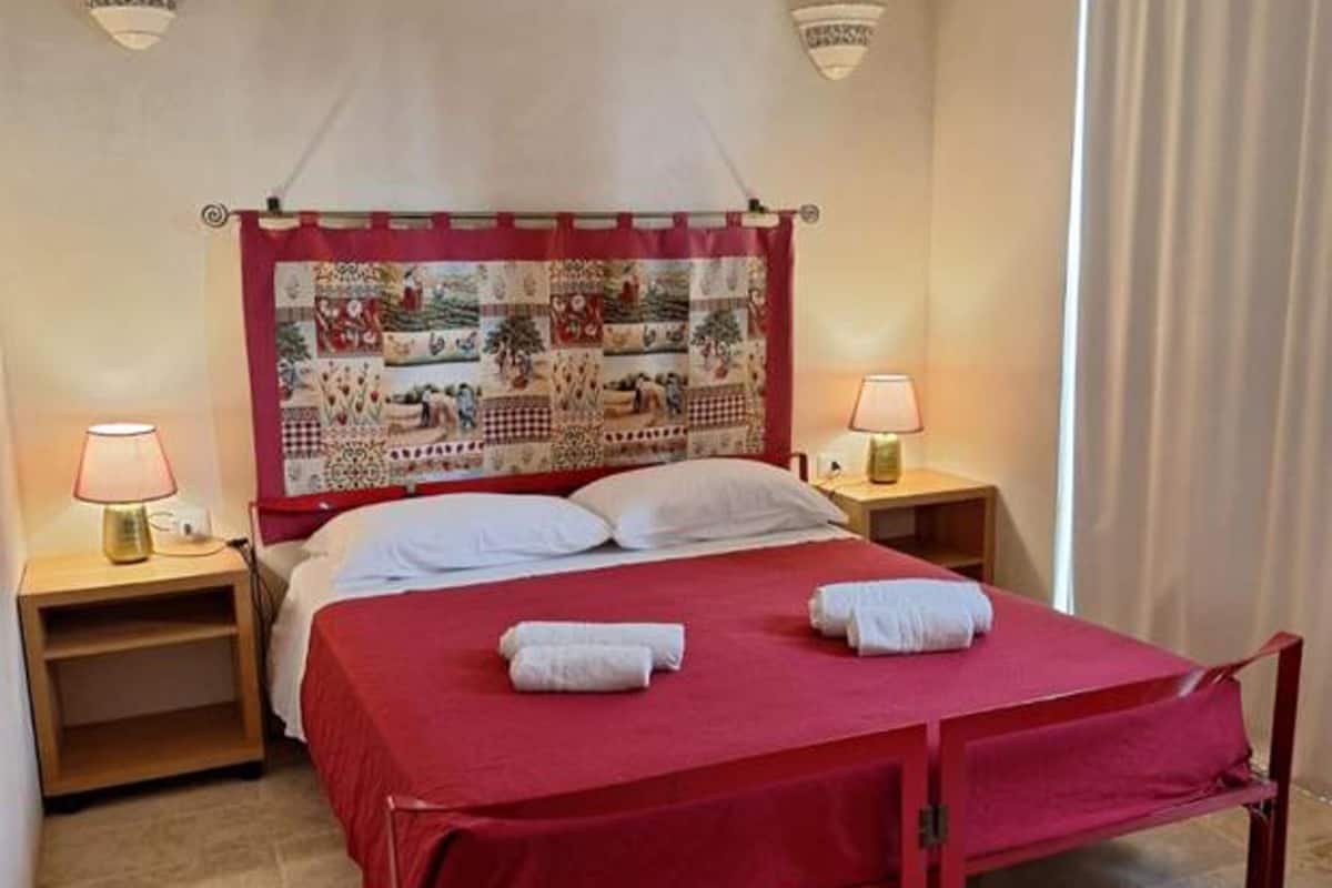 A bed with a red bedspread, two bedside lamps, a quilted headboard, and an air conditioning unit on the wall.