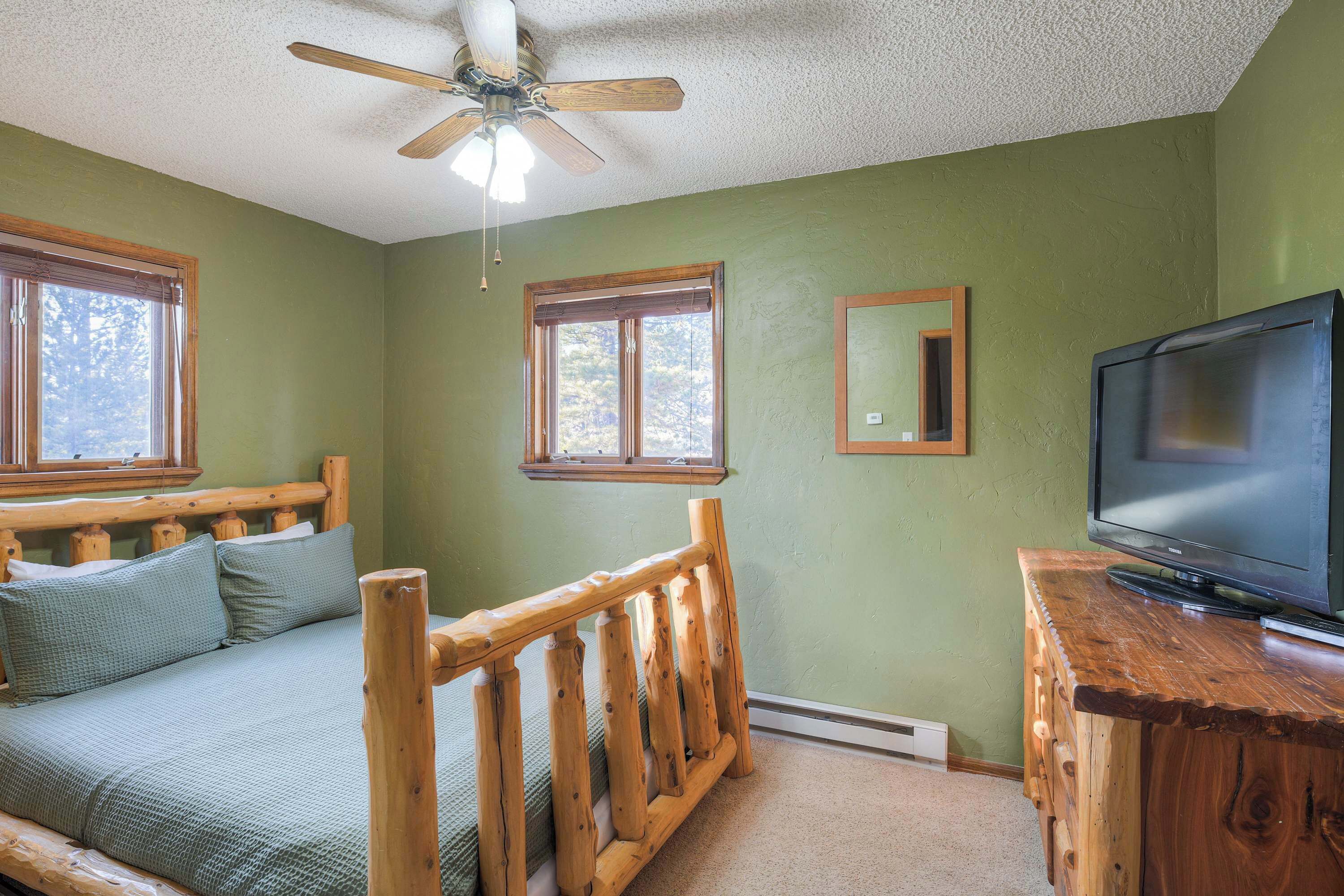 A bedroom with a wooden bed, a ceiling fan, a television on a wooden dresser, and two windows.