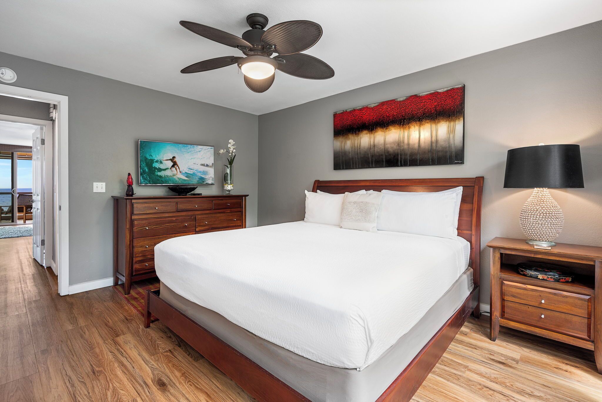 A bedroom with a large bed, a ceiling fan, a painting, a lamp, and a TV on a wooden dresser.