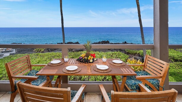 A wooden table set for four with a fruit bowl centerpiece, overlooking a beach with the ocean and palm trees. - Oceanfront - Amazing Views - Completely Renovated - Vrkcv (Kailua-Kona)