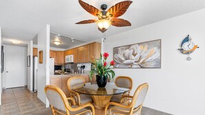 A dining area with a glass-top table surrounded by wicker chairs, a ceiling fan, a potted plant with flowers, and a clock on the wall. - Ocean Front - Fantastic Location - Walk To Kona Town - Vrkcv (Kailua-Kona)