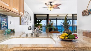 A kitchen with a granite countertop, a stainless steel sink, wooden cabinets, a ceiling fan, and a view of the balcony. - Ocean Front - Fantastic Location - Walk To Kona Town - Vrkcv (Kailua-Kona)
