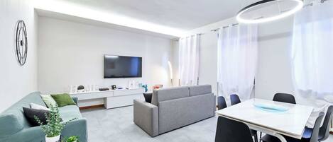 A modern living room with a grey sofa, a dining table with chairs, a flat-screen TV mounted on the wall, and a ceiling light fixture.