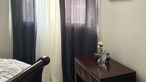 Un dormitorio con una cama, una mesita de noche de madera, un cuadro en la pared y una ventana con cortinas oscuras.