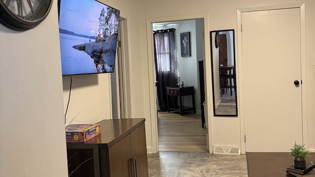 Una sala de estar con televisor de pantalla plana, un mueble de madera oscura, una mesita con una planta y una puerta que da a otra habitación.