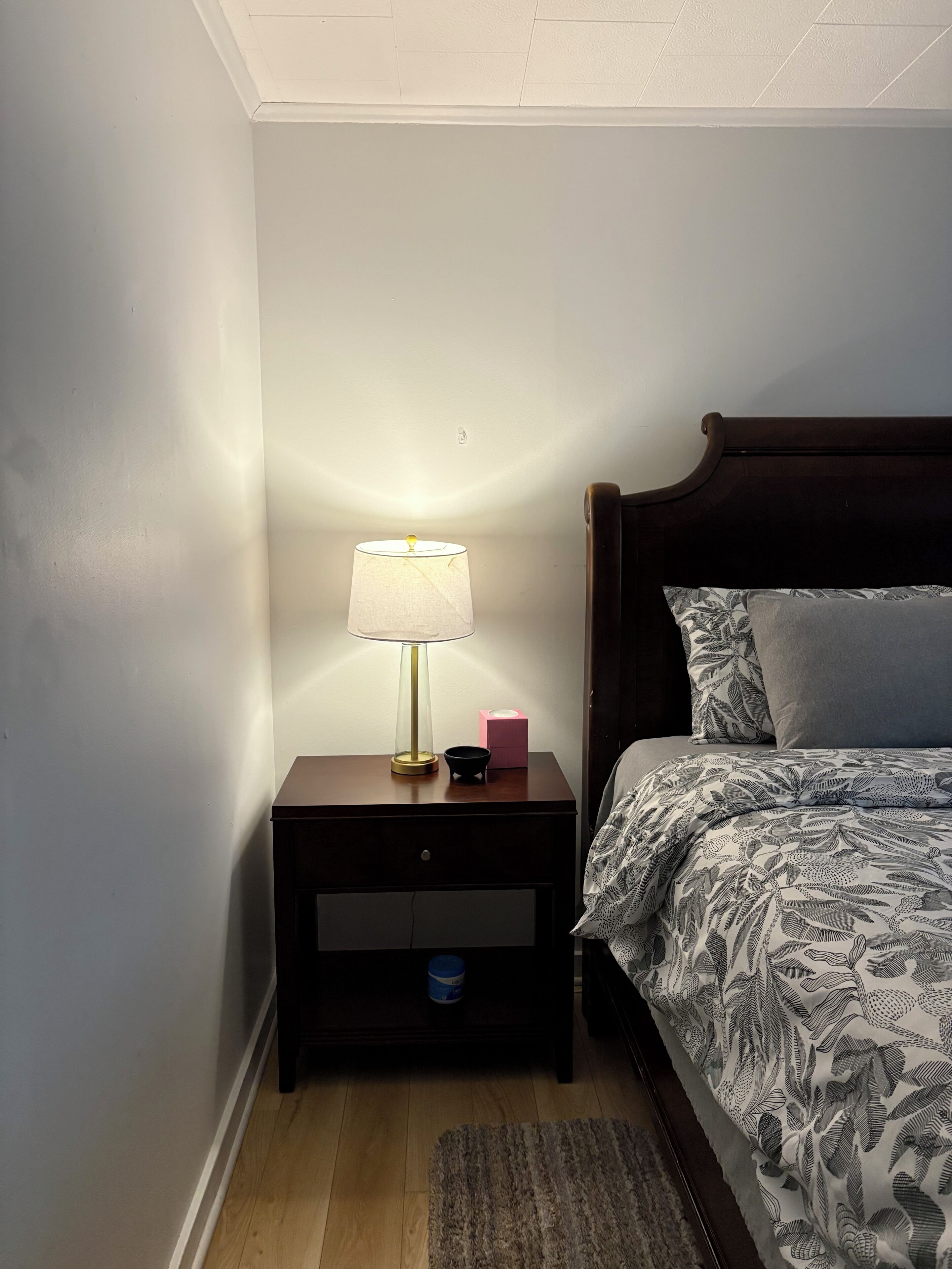 A bedroom with a wooden nightstand, a bedside lamp, a pink tissue box, and a bed with patterned bedding.