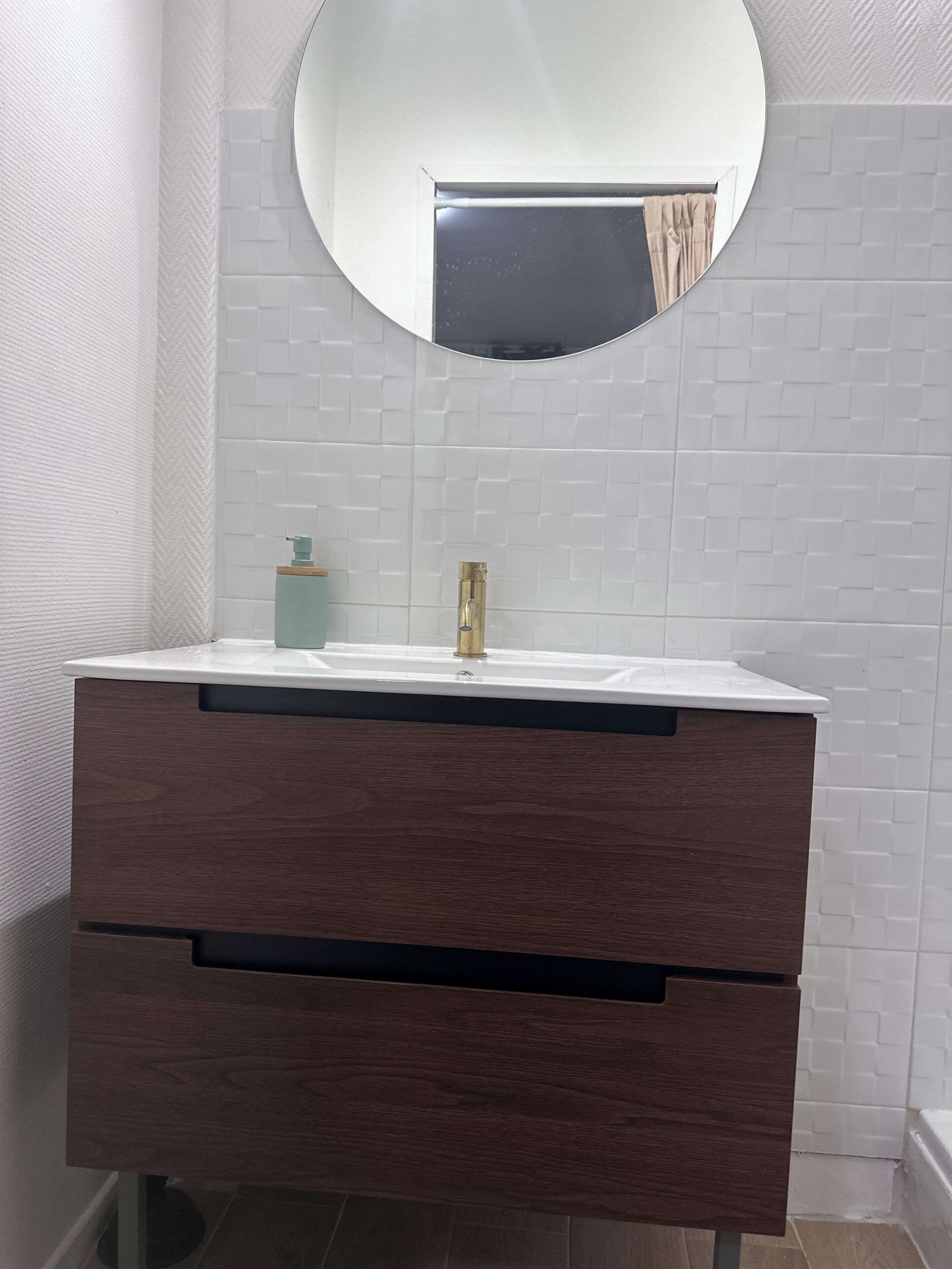 Une salle de bain avec un mur recouvert de carreaux blancs, un miroir rond, un lavabo blanc et un meuble-lavabo en bois doté de deux tiroirs.