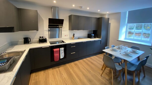 A modern kitchen with dark cabinetry, a central island, and a dining area with a table and chairs. - The Square Apartments Flat 3 (Swindon)