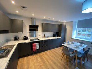 A modern kitchen with dark cabinetry, a central island, and a dining area with a table and chairs. - The Square Apartments Flat 3 (Swindon)