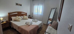 Ein ordentlich bezogenes Bett mit Kissen und Decke, ein Nachttisch mit Lampe, einem Spiegel und ein Fenster mit Vorhängen.