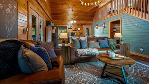A cozy cabin interior with a wooden ceiling, a staircase, a living room with leather sofas, a coffee table, and a kitchen area visible through an open door. - Fresh Design! Creekside Cabin, Hot Tub, Dollywood! (Sevierville)