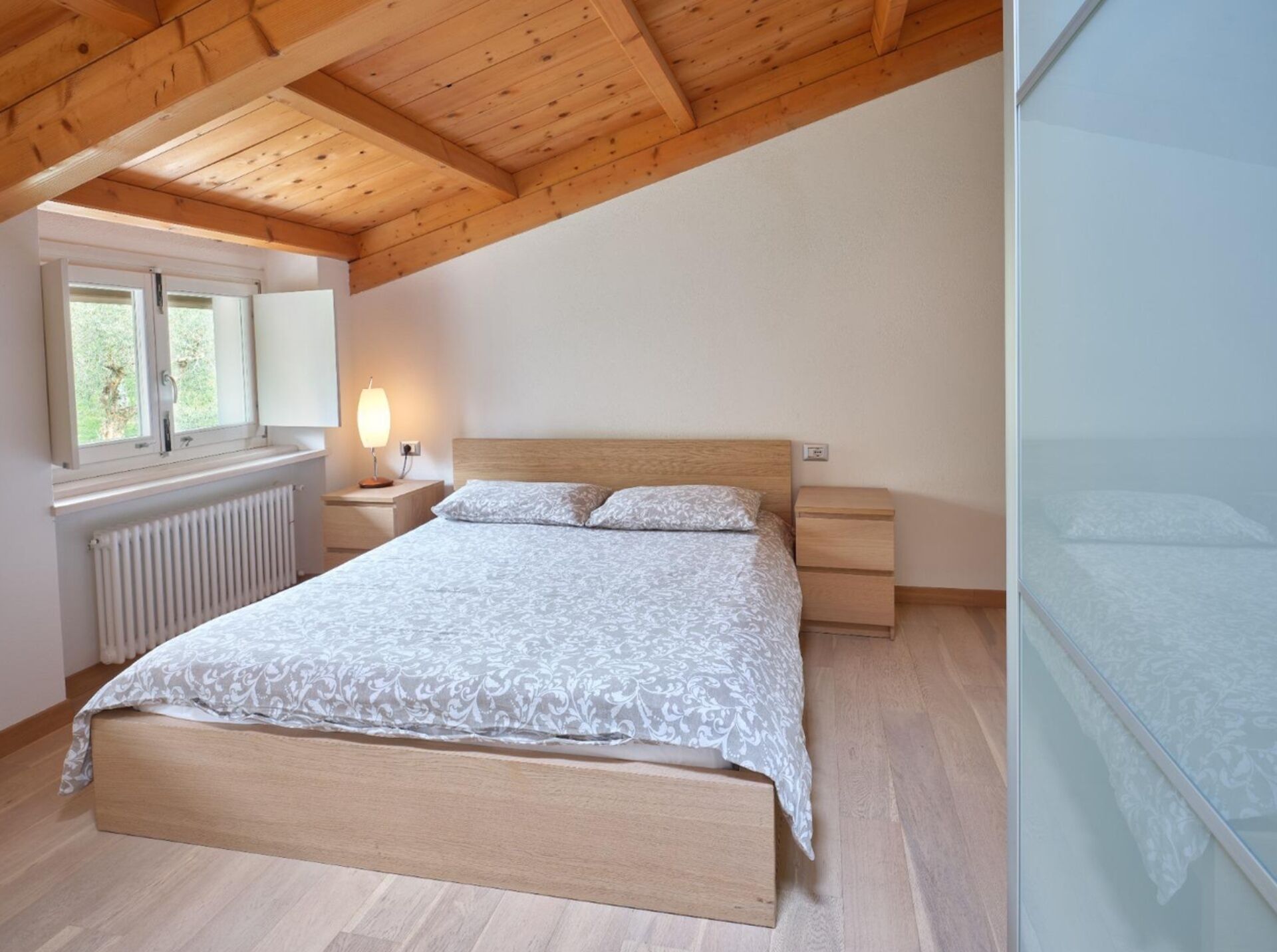 A bedroom with a wooden ceiling, a bed with a patterned bedspread, a nightstand with a lamp, and a window with a view of greenery.