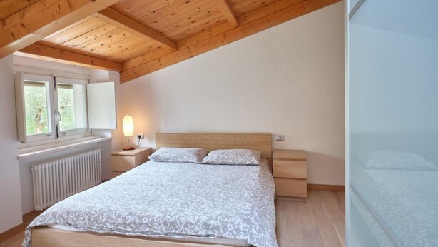 A bedroom with a wooden ceiling, a bed with a patterned bedspread, a nightstand with a lamp, and a window with a view of greenery. - Apartment for 6 people (RIVOLI VERONESE)