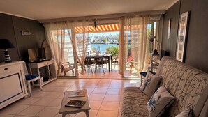A living room with a sofa, a table, a TV, and a view of a pool and buildings. - Marina Giulina harbor view with jetty (Le Grau-du-Roi)