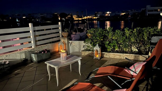 An outdoor seating area with a table and chairs, a railing, and a view of the cityscape at night. - Marina Giulina harbor view with jetty (Le Grau-du-Roi)