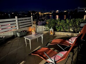 An outdoor seating area with a table and chairs, a railing, and a view of the cityscape at night. - Marina Giulina harbor view with jetty (Le Grau-du-Roi)