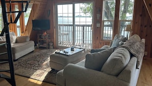 A cozy cabin interior with a large window overlooking a lake, a sofa, a coffee table, and a TV mounted on the wall.