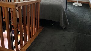 A bedroom with a sloped ceiling, a bed with a grey bedspread, a wooden crib, a standing fan, and a window with a view of trees.