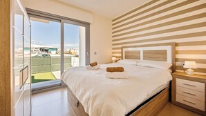 A modern bedroom with a large bed, a wooden headboard, and a view of a cityscape through a sliding glass door. - Casa d'Oro neart Vistabella Golf (Orihuela)