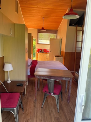 A compact room with a bed, a small kitchenette, a dining table with chairs, and a red pendant light.