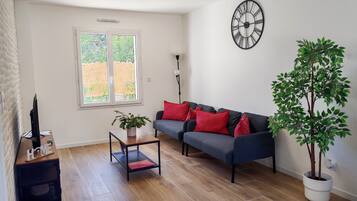 A living room with a sofa, a coffee table, a potted plant, a wall clock, and a window with a view of greenery.