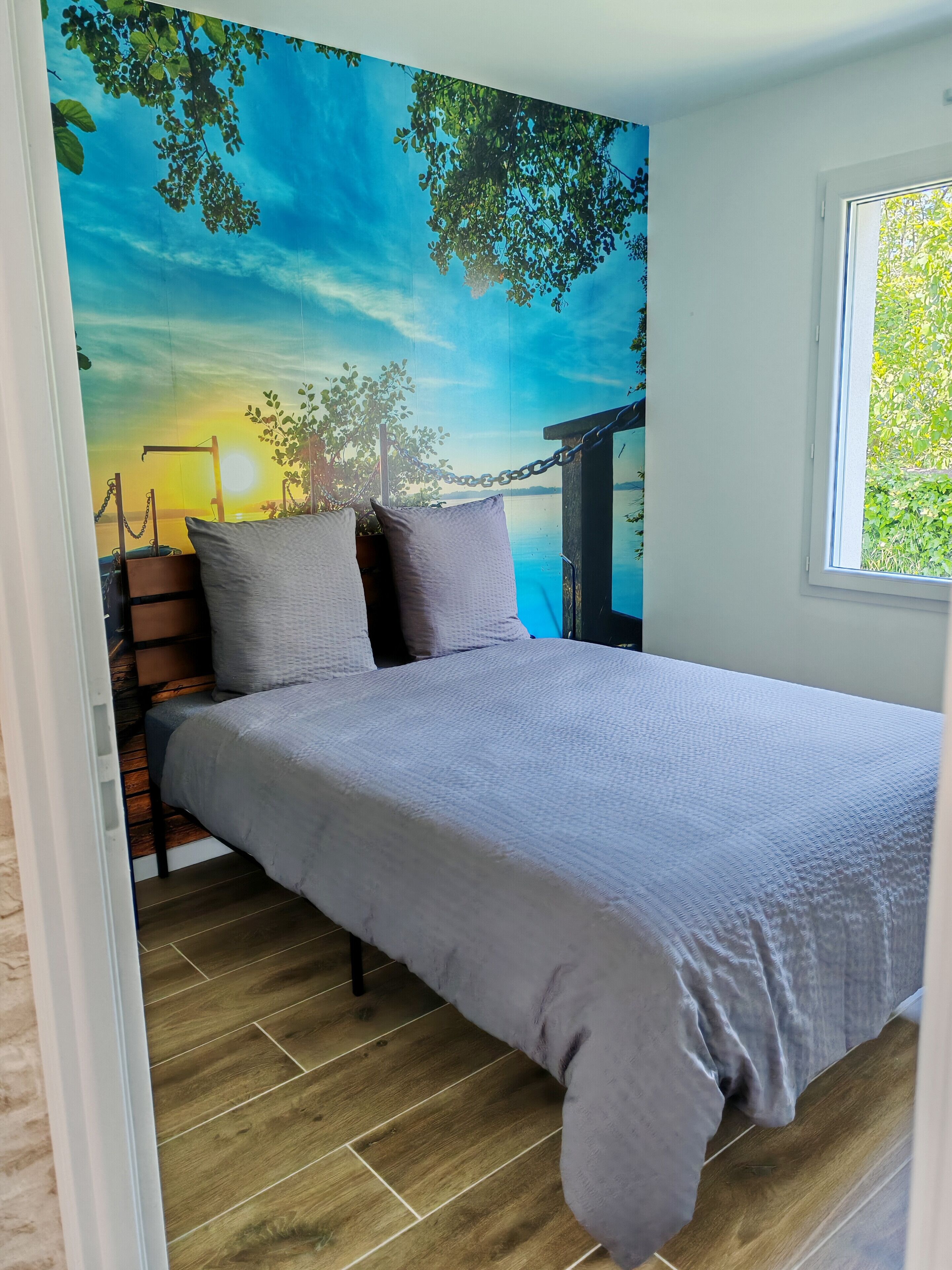 Une chambre à coucher avec un lit, des oreillers, un plancher en bois et une fresque représentant un coucher de soleil.