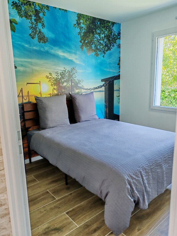 A bedroom with a bed, pillows, a wooden floor, and a mural of a sunset scene.