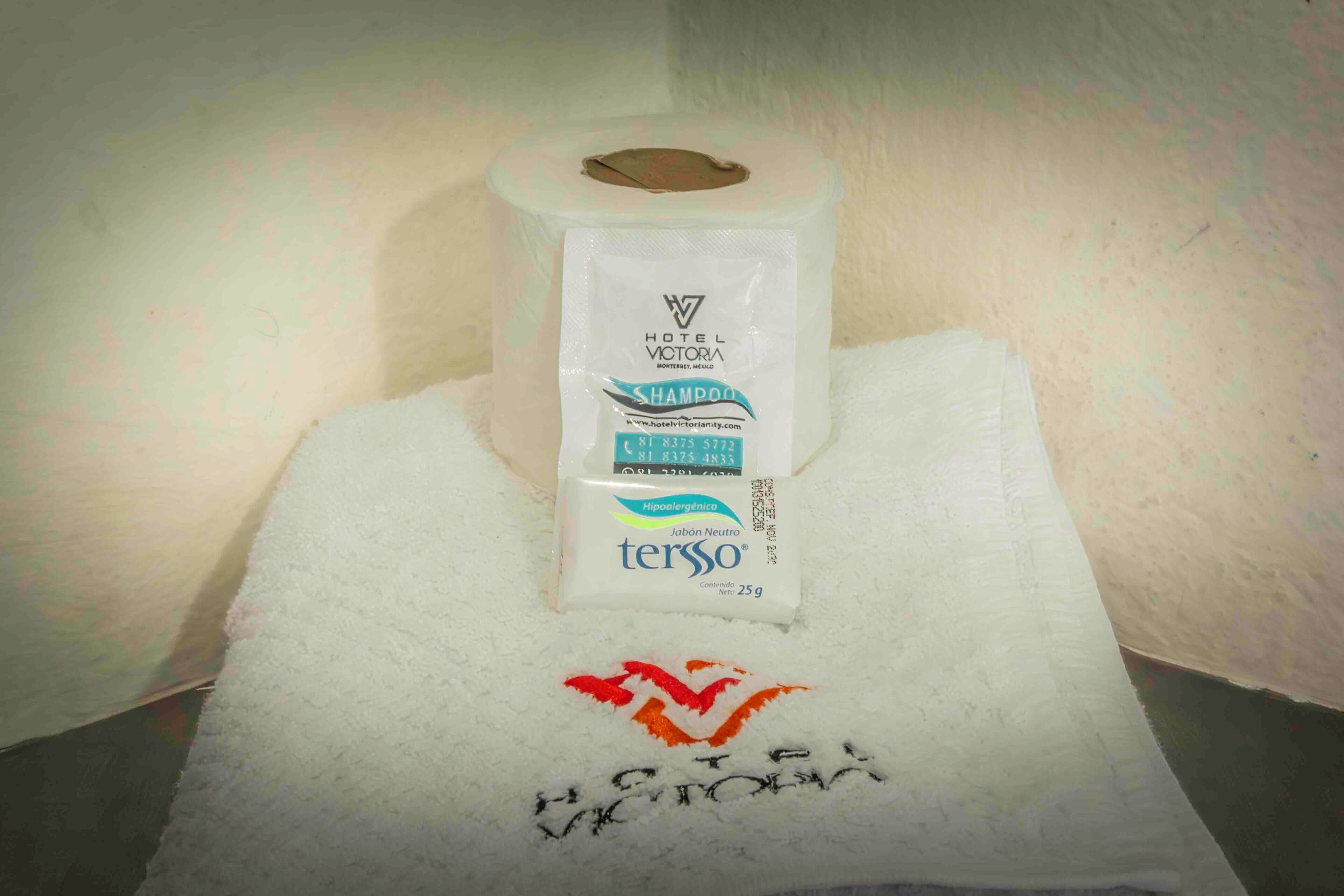 A hotel bathroom with a white towel, a roll of Hotel Victoria shampoo, and a towel with Hotel Victoria embroidered on it.