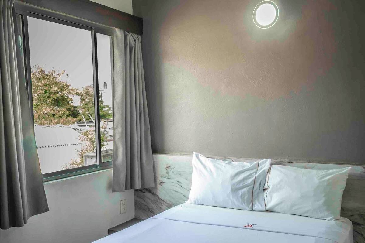 Une chambre à coucher avec un lit, des rideaux, un climatiseur, une fenêtre et un luminaire fixé au mur.