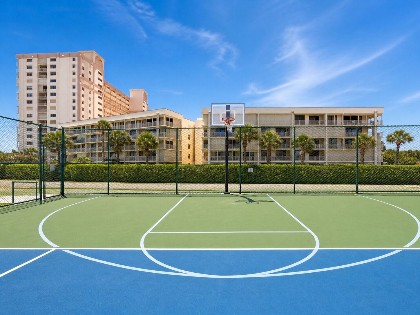 An outdoor basketball court with a net, surrounded by a fence, and buildings in the background.