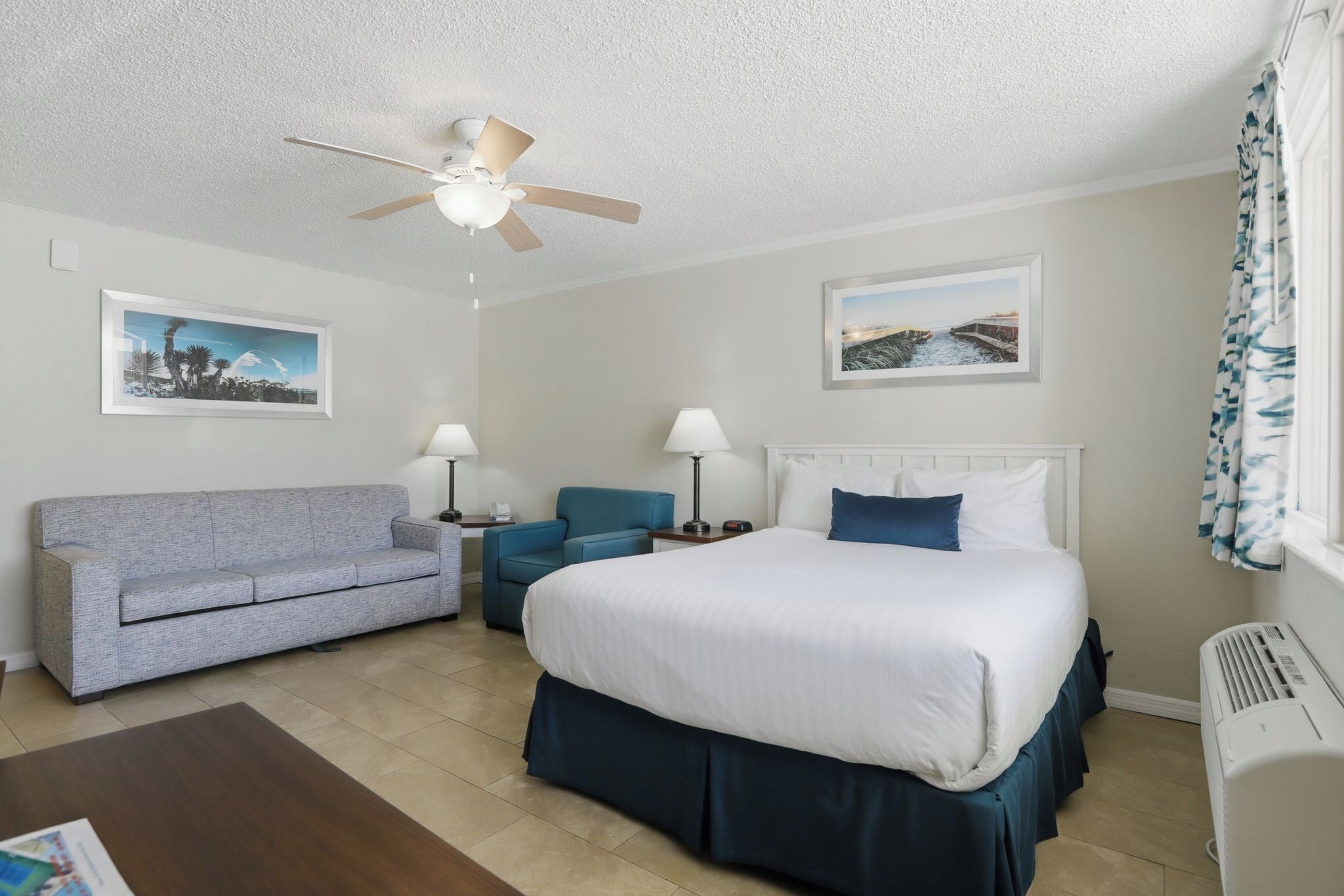 A hotel room with a bed, a sofa, a chair, two lamps, ceiling fan, and two framed pictures on the walls.