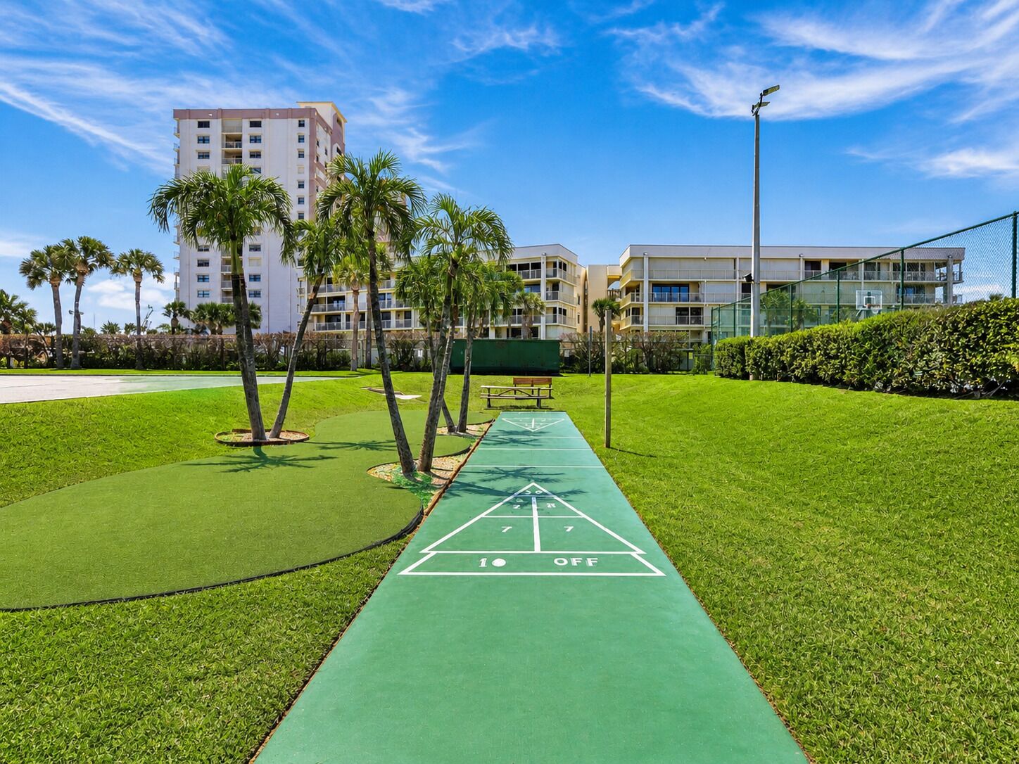 Una cancha de shuffleboard verde al aire libre, cielo despejado, palmeras y un edificio en el fondo.