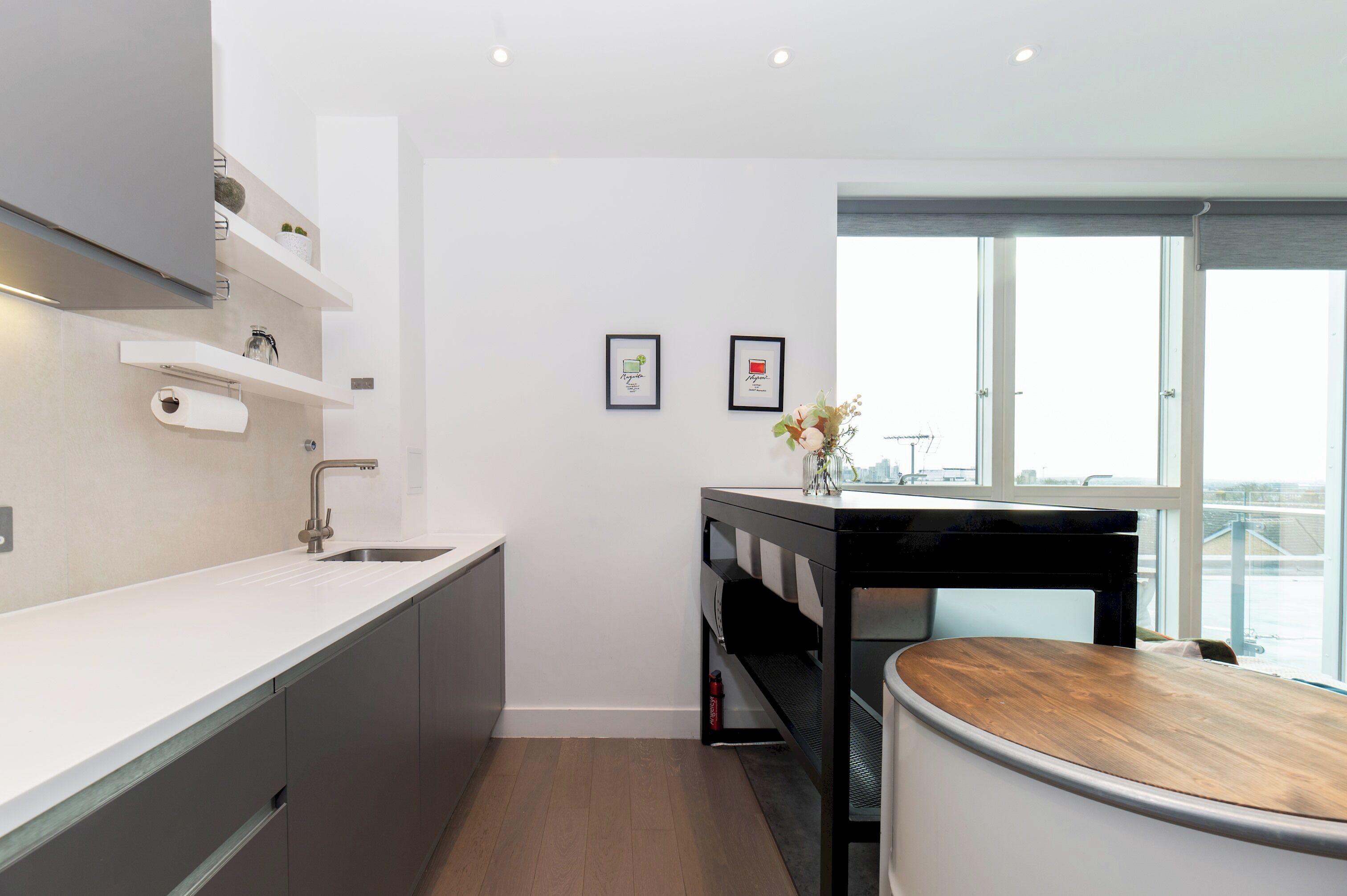 A modern kitchen with a white countertop, grey cabinets, and a black and white bar area. There are two framed pictures on the wall and a window providing a view of the city.