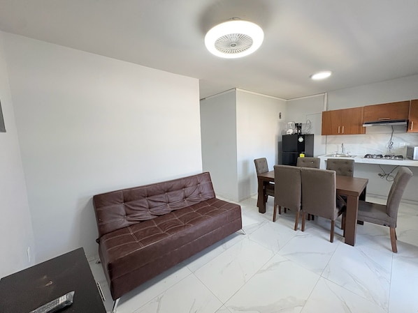 A compact living space with a brown leather sofa, a dining table with chairs, a kitchenette, and a ceiling light fixture.