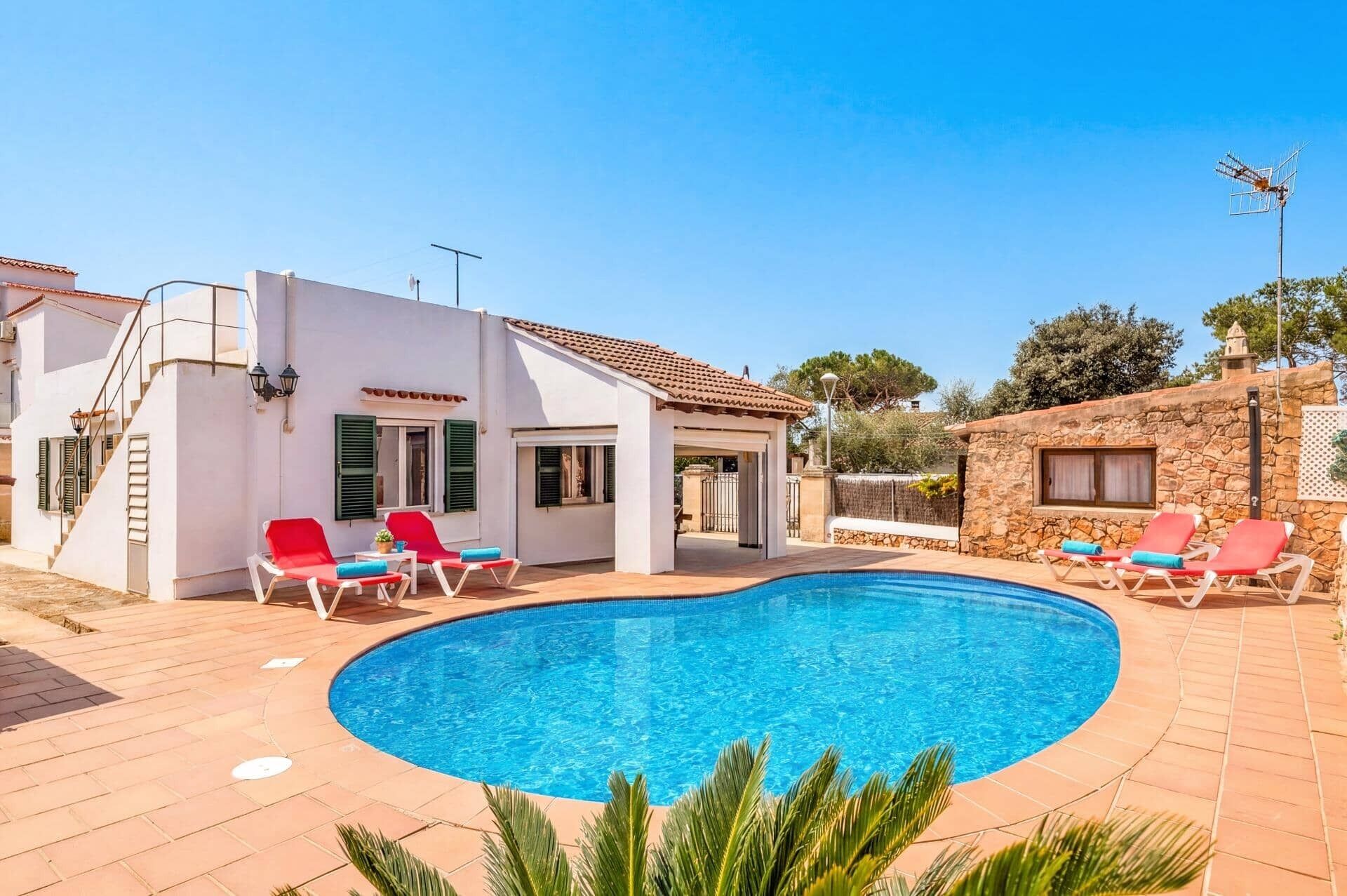 Un espace aménagé au bord de la piscine, avec une piscine, des chaises longues rouges et un bâtiment blanc aux volets verts.