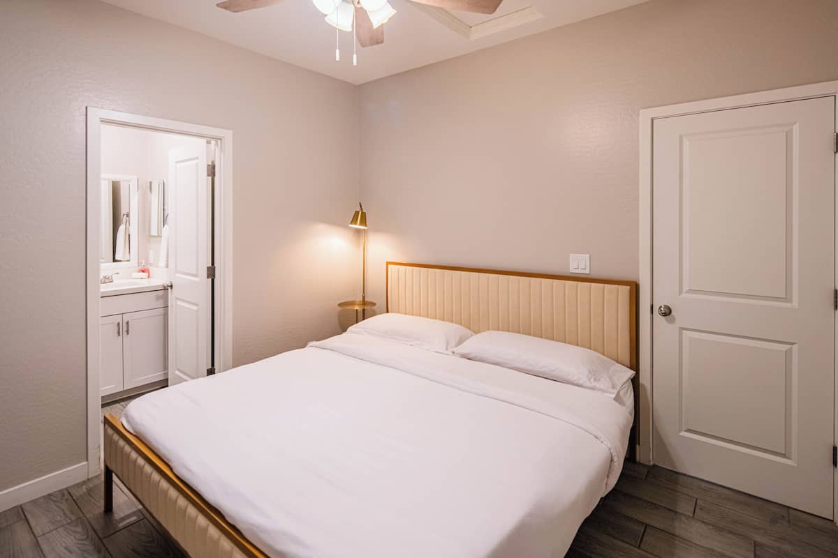 A bedroom with a bed, a nightstand with a lamp, a door, and a bathroom with a mirror and sink.