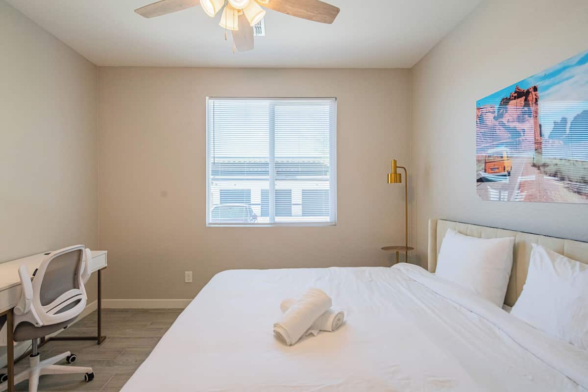 A hotel room with a large bed, a desk with a chair, a ceiling fan, a window with blinds, and a painting on the wall.