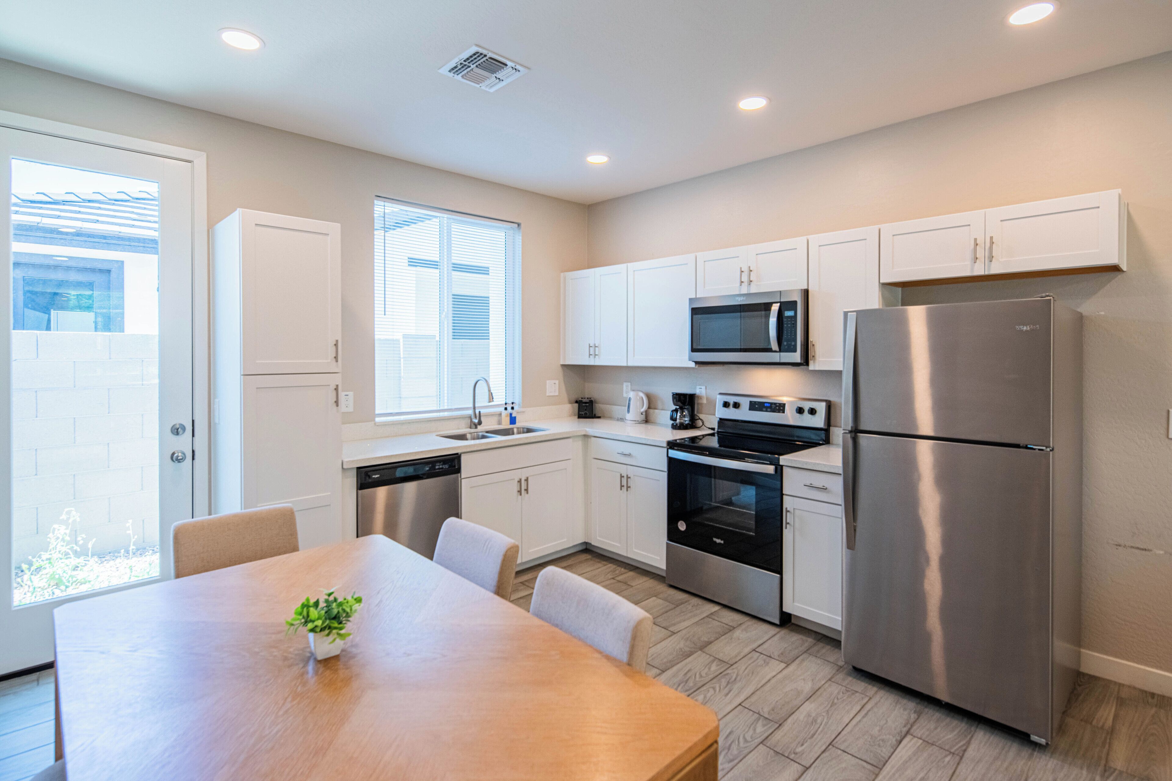 A modern kitchen with white cabinets, stainless steel appliances, a wooden dining table, and a potted plant.