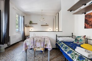 Une pièce comprenant un lit, une table avec une chaise, un radiateur, une fenêtre avec des rideaux et une étagère fixée au mur, sur laquelle se trouve une plante.