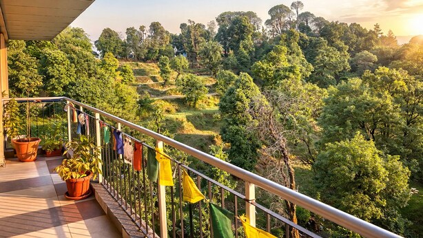 Uma varanda com vasos de plantas e bandeiras, com vista para uma paisagem verdejante com campos em socalcos e árvores densas.