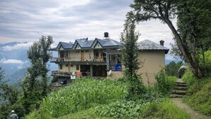 Uma casa de dois andares com varanda, cercada por vegetação exuberante e montanhas ao fundo.