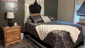 Un dormitorio con cama, mesita de noche, lámpara, ventilador de techo y ventana con cortinas.
