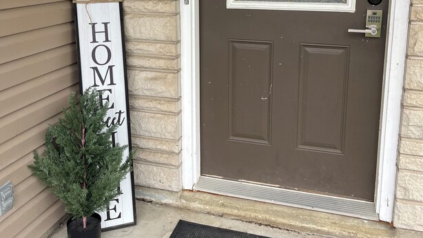 Una puerta principal marrón con un panel de vidrio y un cartel de bienvenida. Un letrero que dice 'HOME' está colgado en la pared a la izquierda.