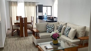 A modern living room with a glass coffee table, a sofa with patterned cushions, a dining area with a wooden table and chairs, and a kitchen in the background.