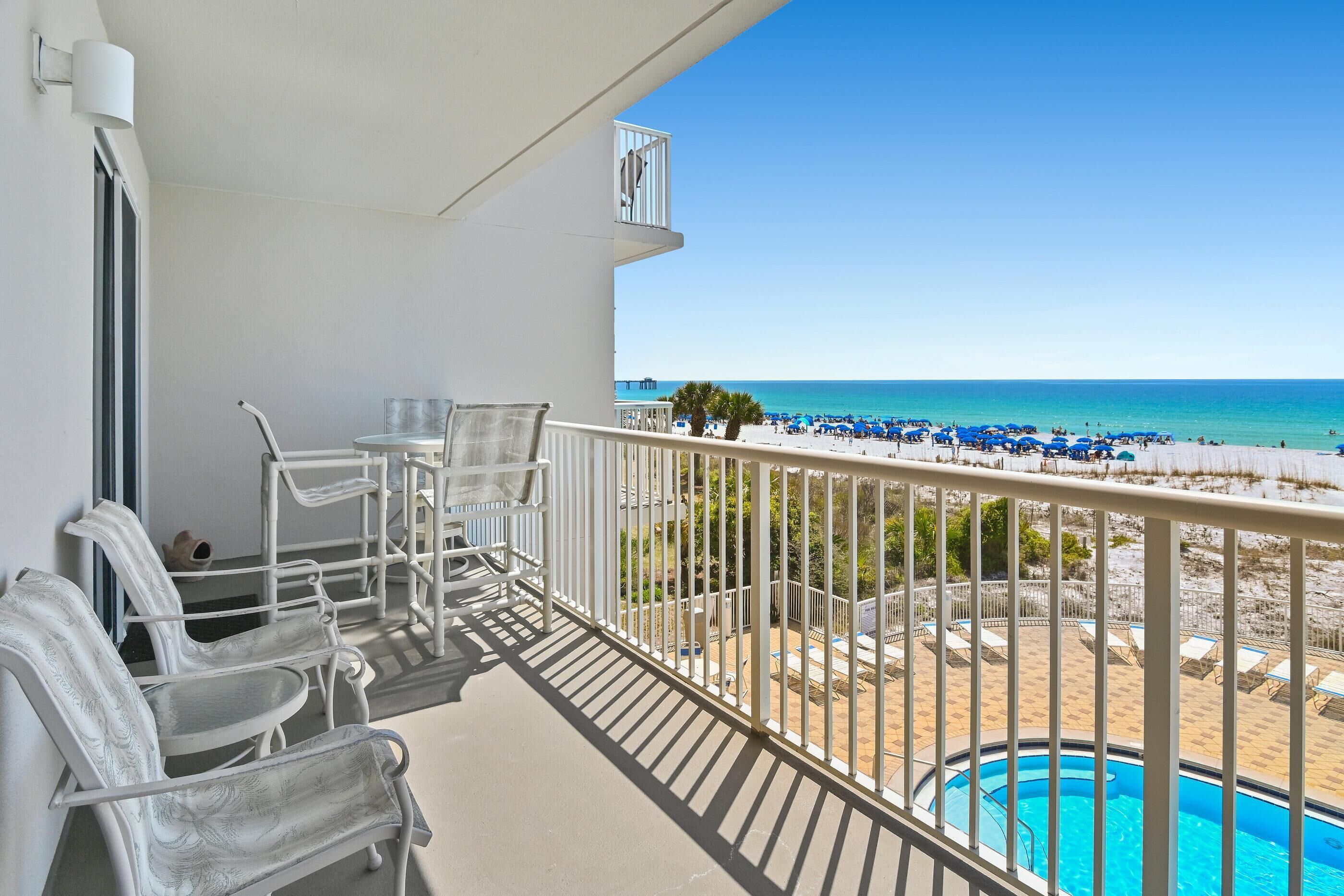 A balcony with white chairs, a small table, and a view of a beach with blue umbrellas and a pool.