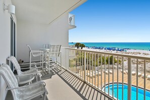 A balcony with white chairs, a small table, and a view of a beach with blue umbrellas and a pool. - Summer Place #303 (Fort Walton Beach)