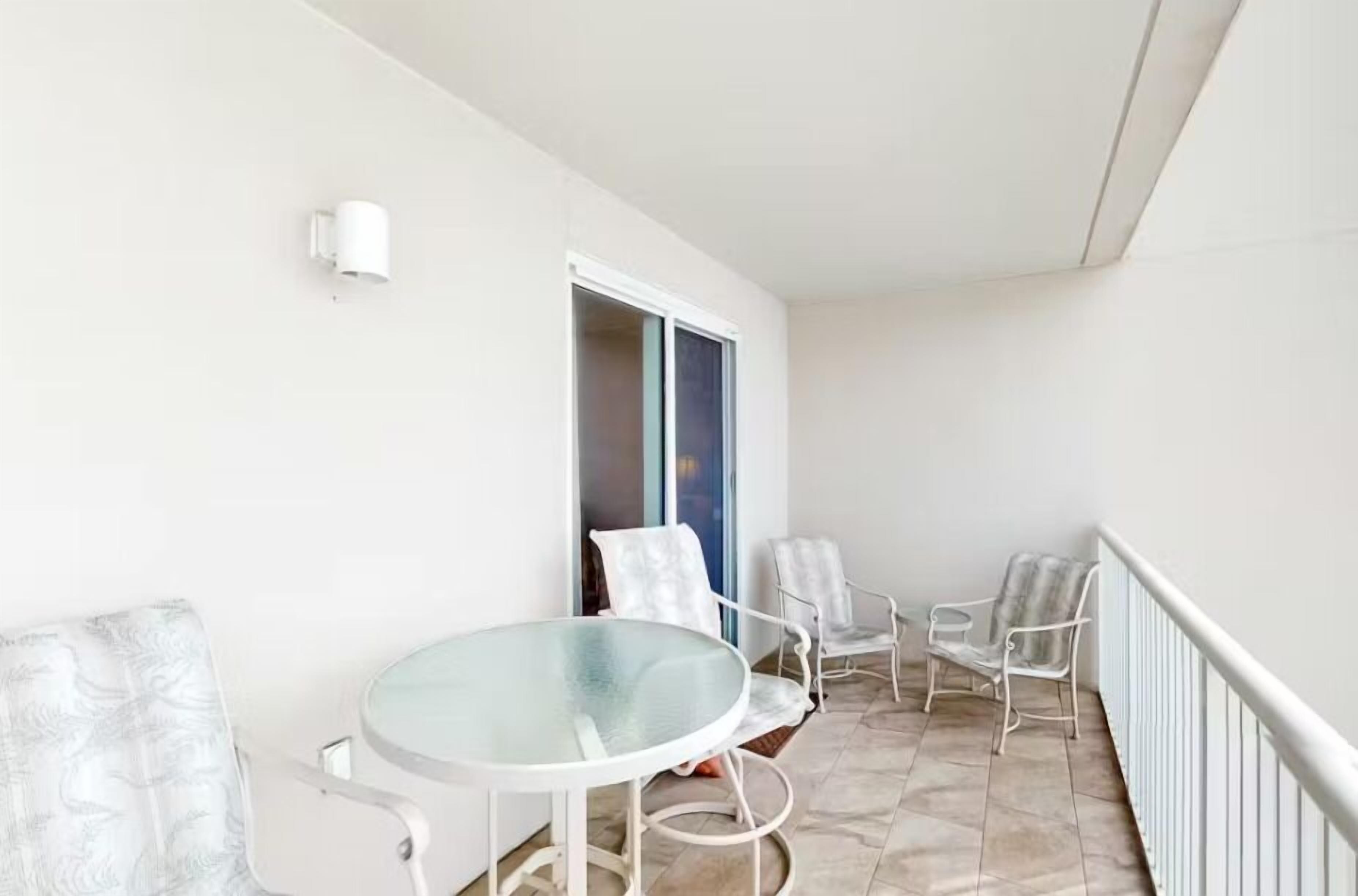 A balcony with a glass table and white chairs, a white wall, and a window with a view.